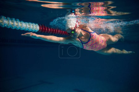 Foto de Coordination of movements. Professional female swimmer in cap and goggles in motion, training in swimming pool indoor. Underwater view. Sport, endurance, competition, energy, healthy lifestyle concept - Imagen libre de derechos