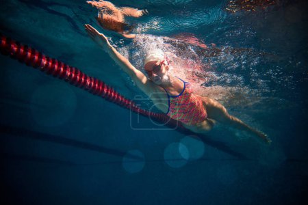 Téléchargez les photos : Front crawl. Young woman, professional swimmer in goggles and cap training, swimming in pool indoors. Underwater view. Concept of sport, endurance, competition, energy, healthy lifestyle - en image libre de droit