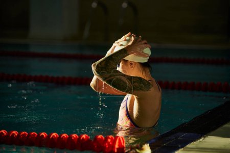 Téléchargez les photos : Preparing to swim. Young woman, professional female swimming athlete in cap and goggles posing in pool indoor. Concept of sport, endurance, competition, energy, healthy lifestyle, power - en image libre de droit