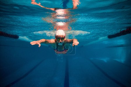 Foto de Fast and strong. Professional female swimmer in cap and goggles in motion, training in swimming pool indoor. Underwater view. Concept of sport, endurance, competition, energy, healthy lifestyle - Imagen libre de derechos