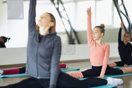 Photo for Gymnastics warm-up. Young woman, sports coach training stretching exercises with little girls, kids indoors. Flexibility. Concept of sportive lifestyle, childhood, education, professional sport. - Royalty Free Image