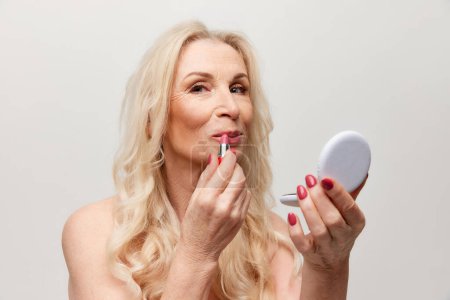 Téléchargez les photos : Beautiful middle-aged woman with well-kept healthy skin applying pink lipstick over grey studio background. Mature old lady. Concept of natural beauty, face skin care, cosmetology and cosmetic, health - en image libre de droit
