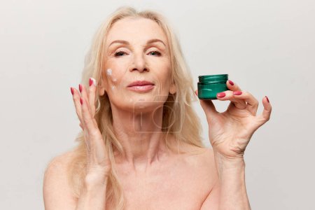 Téléchargez les photos : Attractive middle-aged woman applying moisturizing, nourishing face cream. Mature old woman posing against grey background. Concept of natural beauty, face skin care, cosmetology and cosmetics, health - en image libre de droit