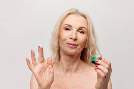 Téléchargez les photos : Everyday skin routine. Beautiful middle-aged woman applying moisturizing face cream. Mature old lady posing on grey background. Concept of natural beauty, face skin care, cosmetology and cosmetics - en image libre de droit