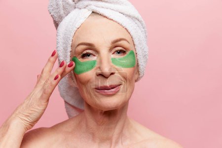 Foto de Close-up of beautiful middle-aged woman with towel on head posing with under eye patches, posing on pink studio background. Concept of natural beauty, face skin care, cosmetology and cosmetics, health - Imagen libre de derechos