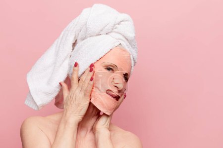 Téléchargez les photos : Beautiful middle-aged woman over pink studio background. After shower face care, applying mask. Mature treatment. Concept of natural beauty, face skin care, cosmetology and cosmetics, health - en image libre de droit