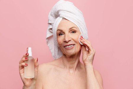 Téléchargez les photos : Hydration and refreshment. Beautiful middle-aged, mature woman applying face toner over pink studio background. Concept of natural beauty, face skin care, cosmetology and cosmetics, health - en image libre de droit
