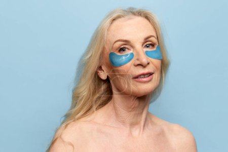 Téléchargez les photos : Beautiful middle-aged woman with well-kept healthy skin posing with under eye patches over blue studio background. Concept of natural beauty, face skin care, cosmetology and cosmetics, health - en image libre de droit