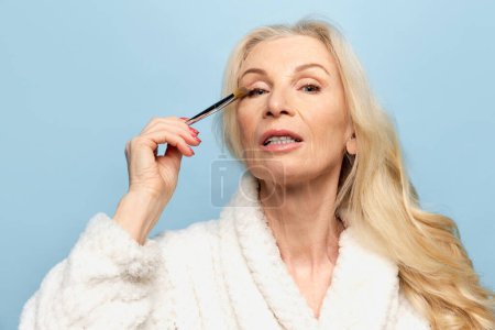 Foto de Beautiful middle-aged, old woman applying eye mascara, getting ready over blue studio background. Mature model. Concept of natural beauty, face skin care, cosmetology and cosmetics, health - Imagen libre de derechos