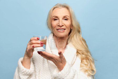 Foto de Importancia de la hidratación. Hermosa mujer de mediana edad sonriendo, posando con un vaso de agua sobre fondo azul del estudio. Concepto de belleza natural, cuidado de la cara, cosmetología y cosmética, salud - Imagen libre de derechos