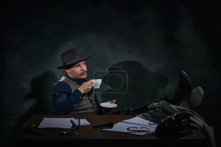 Téléchargez les photos : Professional detective in fedora hat sitting at table with thoughtful face and drinking coffee over dark green vintage background. Inspector. Concept of occupation, character, history. Retro style - en image libre de droit