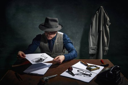 Photo for Professional detective in fedora hat sitting at table and looking at footprint, crime clue over dark green vintage background. Investigation. Concept of occupation, character, history. Retro style - Royalty Free Image