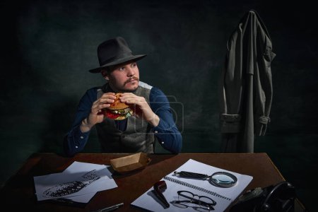 Foto de Detective profesional en sombrero fedora sentado en la mesa y comiendo deliciosa hamburguesa para el almuerzo sobre fondo vintage verde oscuro. Concepto de ocupación, carácter, historia. Estilo retro - Imagen libre de derechos