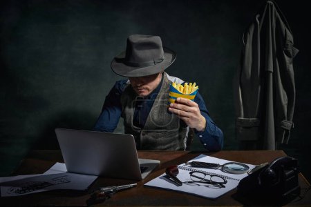 Foto de Professional detective in fedora hat sitting at table, working on laptop and eating fries over dark green vintage background. Concept of occupation, character, history. Retro style - Imagen libre de derechos