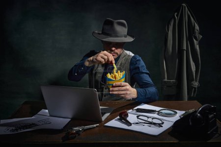 Téléchargez les photos : Professional detective in fedora hat sitting at table, working on laptop and eating fries over dark green vintage background. Busy man. Concept of occupation, character, history. Retro style - en image libre de droit