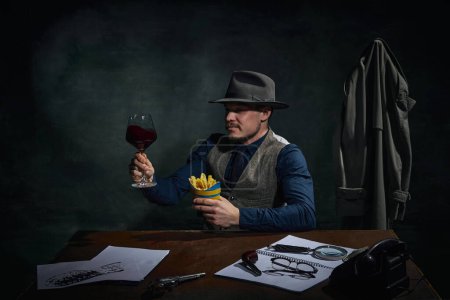 Photo for Professional detective in fedora hat sitting at table, drinking red wine and eating fries over dark green vintage background. Concept of occupation, character, history. Retro style - Royalty Free Image