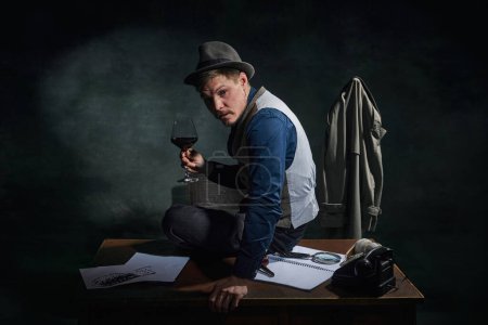 Téléchargez les photos : Professional detective in fedora hat and classical clothes sitting on table with glass of red wine over dark green vintage background. Concept of occupation, character, history. Retro style - en image libre de droit