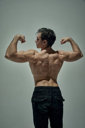 Téléchargez les photos : Strong relief back and biceps. Male model posing shirtless, in pants over pale green studio background. Muscular body shape. Concept of mans beauty, sportive and healthy lifestyle, fashion - en image libre de droit