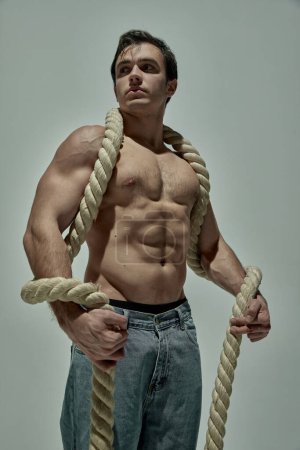 Photo for Handsome young man with strong muscular body shape posing in jeans with rope over grey studio background. Concept of mans beauty, sportive and healthy lifestyle, fashion - Royalty Free Image