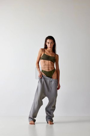 Téléchargez les photos : Full-length portrait of beautiful young girl with slim, fit body posing in underwear and oversized pants on grey studio background. Concept of beauty, weight loss, body-positivity, wellness, body care - en image libre de droit