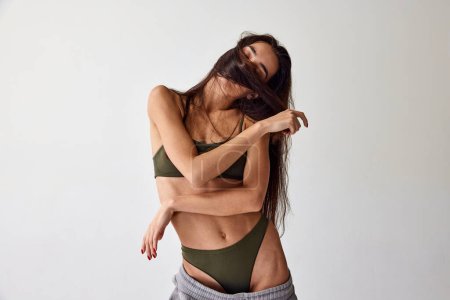 Téléchargez les photos : Body positivity. Tender young brunette girl with fit muscular body poisoning underwear over grey studio background. Concept of natural beauty, femininity, wellness, body care and fitness - en image libre de droit