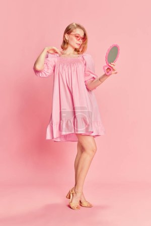 Foto de Retrato de cuerpo entero de una tierna y hermosa joven posando con un elegante vestido rosa, y gafas de sol, mirando en el espejo sobre el fondo del estudio rosa. Concepto de belleza, emociones, moda, estilo de vida, juventud - Imagen libre de derechos