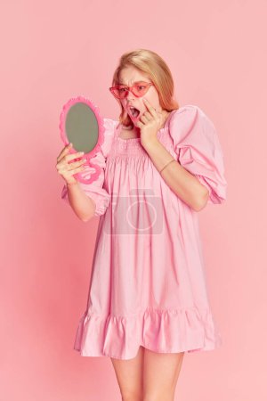 Téléchargez les photos : Finding wrinkles. Emotional young girl in pink dress looking in mirror with expressive face over pink studio background. Concept of beauty, emotions, fashion, lifestyle and youth culture. - en image libre de droit