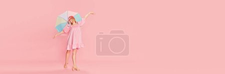 Téléchargez les photos : Happy beautiful young girl in pink dress, sunglasses and colorful umbrella posing over pink studio background. Concept of beauty, emotions, fashion, lifestyle, youth culture. Banner. Copy space for ad - en image libre de droit