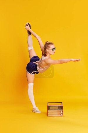 Foto de Full-length image of beautiful slim young girl in sunglasses doing stretching exercises, training over yellow studio background. Concept of beauty, sportive lifestyle, hobby, emotions. Ad - Imagen libre de derechos
