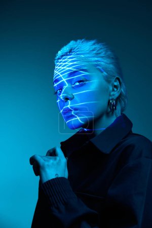Foto de Tecnologías modernas. Retrato de una joven rubia con rayas de neón en la cara posando sobre fondo oscuro en luces de neón azules. Concepto de arte, estilo moderno, cyberpunk, futurismo y creatividad - Imagen libre de derechos