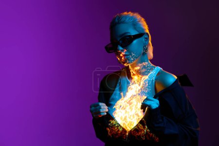 Téléchargez les photos : Brûlant à l'intérieur. Portrait de jeune fille blonde avec feu numérique sur le corps posant sur fond violet dans des néons bleus. Concept d'art, style moderne, cyberpunk, futurisme et créativité - en image libre de droit