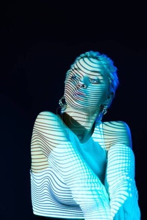 Photo for Futuristic. Portrait of young blonde girl with neon stripes on face posing over dark background in blue neon lights. Concept of art, modern style, cyberpunk, futurism and creativity - Royalty Free Image