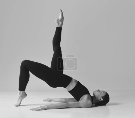 Téléchargez les photos : Slim fit body. Black and white photography. Young girl doing stretching, press exercises, training over studio background. Concept of sport, body care, beauty, fitness, active lifestyle. Ad - en image libre de droit