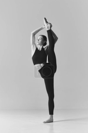 Foto de Fotografía en blanco y negro. Joven chica flexible haciendo ejercicios de estiramiento, de pie sobre cordel sobre fondo del estudio. Concepto de deporte, cuidado corporal, belleza, fitness, estilo de vida activo. Anuncio - Imagen libre de derechos