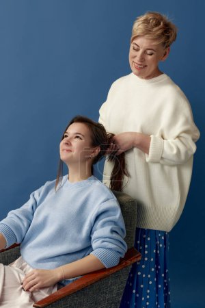 Foto de Retrato de una mujer feliz de mediana edad haciendo peinado a su hija pequeña, posando sobre fondo de estudio azul. Concepto de maternidad, familia, día de la madre, amor, emociones, relación - Imagen libre de derechos
