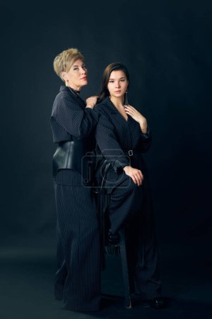 Foto de Full-length portrait of beautiful middle-aged woman, mother posing with her young daughter over dark studio background. Concept of motherhood, family, mothers day, love, emotions, relationship - Imagen libre de derechos