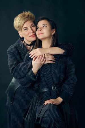 Photo for Hugging. Beautiful middle-aged woman, mother with young girl posing in stylish suits over dark studio background. Concept of motherhood, family, mothers day, love, emotions, relationship - Royalty Free Image