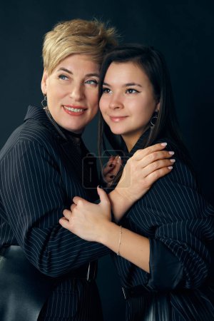 Téléchargez les photos : Happy mother and daughter. Beautiful middle-aged woman posing with young girl over dark studio background. Concept of motherhood, family, mothers day, love, emotions, relationship - en image libre de droit