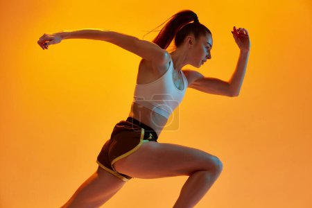 Foto de Acelera. Chica joven, atleta corredor profesional en entrenamiento uniforme, corriendo sobre fondo de estudio naranja en luz de neón. Concepto de estilo de vida deportivo, salud, resistencia, acción y movimiento. Anuncio - Imagen libre de derechos