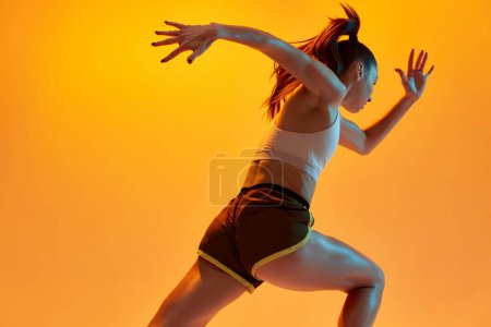 Téléchargez les photos : Speed, strength. Young girl, professional athlete, runner in motion, training over orange studio background in neon light. Concept of sportive lifestyle, health, endurance, action and motion. Ad - en image libre de droit