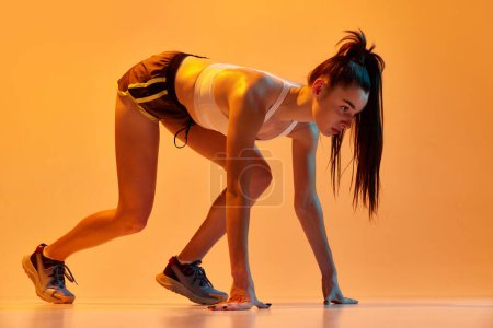 Foto de Listo para correr. Joven atleta corriendo en posición de inicio, entrenando sobre fondo de estudio naranja en luz de neón. Concepto de estilo de vida deportivo, salud, resistencia, acción y movimiento. Anuncio - Imagen libre de derechos