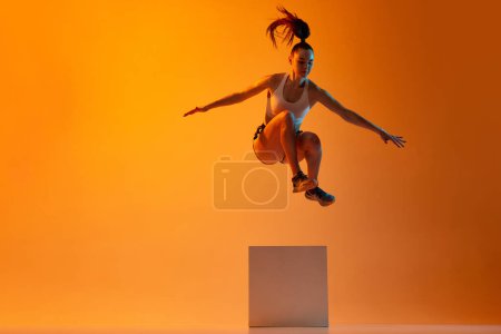 Foto de Workout. Young sportive girl, athlete training, jumping over block against orange studio background in neon light. Concept of sportive lifestyle, health, endurance, action and motion. Ad - Imagen libre de derechos