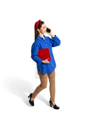 Foto de Young employee, woman in official clothes talking on phone on her way, posing over white studio background. Isometric view. Concept of career development, employment, business, job. Ad - Imagen libre de derechos