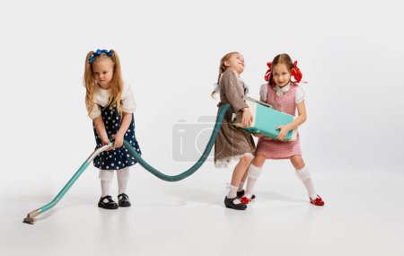 Téléchargez les photos : Petites belles filles, enfants en robes rétro jouant avec l'aspirateur sur fond de studio gris. Concept d'enfance, jeu, amitié, activité, temps libre, style rétro, mode. - en image libre de droit