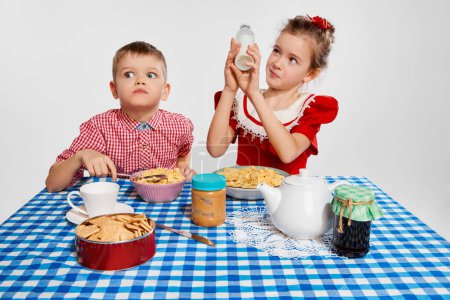 Téléchargez les photos : Emotional kids. Little boy and girl, brother and sister having breakfast together against grey studio background. Concept of childhood, game, friendship, activity, leisure time, retro style, fashion. - en image libre de droit