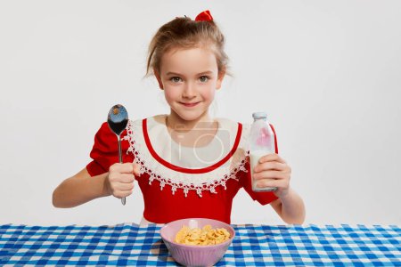 Téléchargez les photos : Cute beautiful little girl having yummy healthy breakfast with cereal, posing against grey studio background. Concept of childhood, game, friendship, activity, leisure time, retro style, fashion. - en image libre de droit