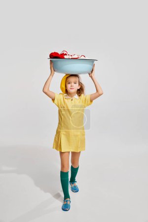 Foto de Beautiful little girl, kid in retro yellow dress doing laundry, posing against grey studio background. Concept of childhood, game, emotions, activity, leisure time, retro style, fashion. - Imagen libre de derechos
