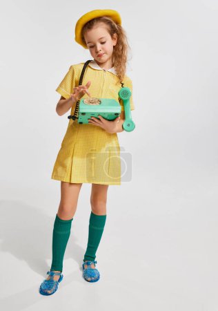 Foto de Beautiful little girl, kid in retro yellow dress playiong with phone, posing against grey studio background. Concept of childhood, game, emotions, activity, leisure time, retro style, fashion. - Imagen libre de derechos