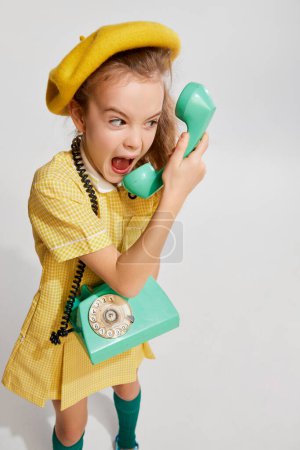 Photo for Beautiful little girl, kid in retro yellow dress emotionally talking on phone, posing against grey studio background. Concept of childhood, game, emotions, activity, leisure time, retro style, fashion - Royalty Free Image
