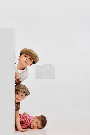 Foto de Tres niños pequeños, chicos espiando esquina, jugando contra el fondo gris del estudio. Concepto de infancia, juego, amistad, actividad, tiempo libre, estilo retro, moda. - Imagen libre de derechos